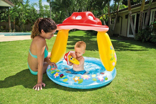 Een kindje speelt in een blauw met geel en rood opblaasbaar kinderzwembad van Intex in de vorm van een paddestoel terwijl zijn moeder naast het bad in de tuin zit