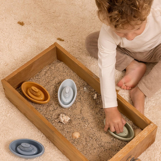 Een kindje speelt met vier bad bootjes van Little Dutch. De bootjes hebben de kleuren blauw, lichtblauw, groen en okergeel. Het kindje speelt in de zandbak