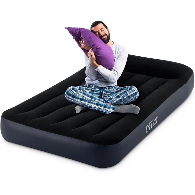 Laad afbeelding in galerijtoner, Een man in pyjama zit in kleermakerszit op een Intex Pillow Rest Classic Eenpersoons Luchtbed, knuffelt een paars kussen en kijkt tevreden.
