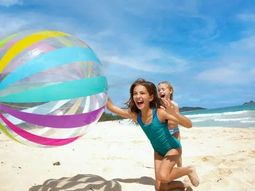 Twee meisjes spelen met een Intex Strandbal Jumbo op een zonnig strand, met de blauwe zee en de lucht achter zich: de perfecte dag met de Intex strandbal bij het strand en zwembad.